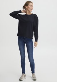 Schwarzer, langärmliger Sweatshirt mit rundem Ausschnitt, kombiniert mit dunkelblauen Skinny-Jeans und grauen Sneakers vor einem hellen Hintergrund.