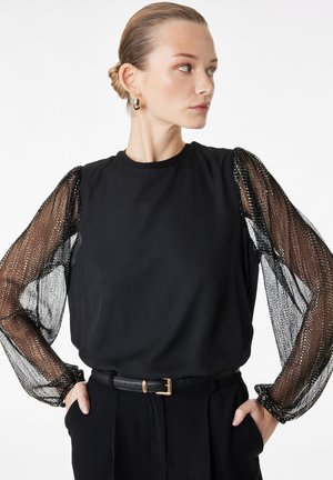 Vrouw in zwarte top met doorschijnende, glinsterende lange mouwen, gouden oorringen en handen in de zakken, kijkend naar de zijkant tegen een witte achtergrond.