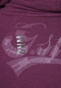 Tessuto viola con un motivo testurizzato, in evidenza un dettaglio testuale "DNM" in argento metallico e un sottile design grafico in tonalità più chiare.