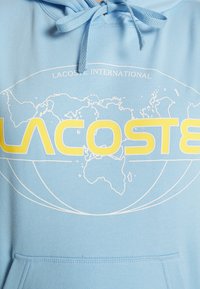 Sweat à capuche bleu clair avec texte jaune "LACOSTE" sur un contour de carte du monde blanc et "LACOSTE INTERNATIONAL" au-dessus, poche avant visible.