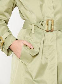 Trench coat verde claro hecho de tela de satén. Presenta una cintura ajustada con un cinturón, un bolsillo grande y puños abotonados. Textura suave.