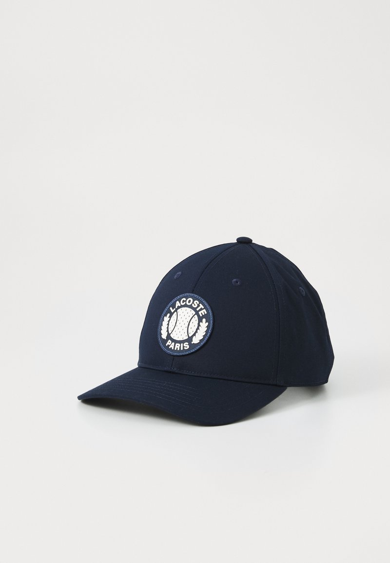 Navy baseball cap made of cotton with a round white patch featuring a tennis motif and "LACOSTE PARIS" text, six-panel design, and curved brim.