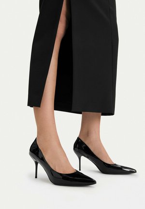 Mujer con zapatos de tacón alto de punta afilada de charol negro y tacones de aguja, y una falda midi negra con una abertura frontal.