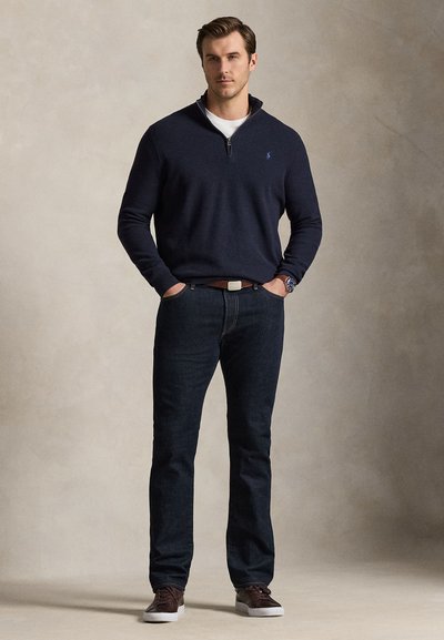 Homme debout, les mains dans les poches, portant un pull bleu marine à fermeture éclair quart, un jean foncé, une ceinture marron, une montre et des baskets marron sur un fond uni.