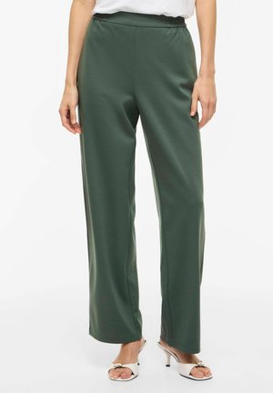 Mørkegrønne bukser med rett snitt, båret av en stående person, kombinert med hvite åpne hælsandaler og en hvit topp stukket inn.