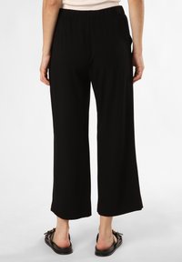 Schwarze, weite Hosen mit einem elastischen Bund und Taschen. Glatte Stofftextur. Kombiniert mit schwarzen Sandalen, die ein Schleifendetail haben.