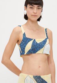 Izgriezta blūze ar zilu pērļu rakstu uz dzeltena auduma, ar platām siksnām un kvadrātveida kakla izgriezumu. Tekstūras akcenti gaismas zilos toņos.