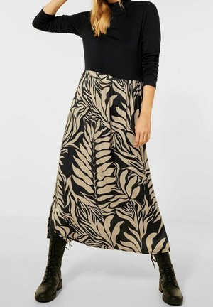 Femme portant un haut noir à manches longues, une jupe midi beige à motifs de feuilles noires et des bottes noires à lacets, debout devant un fond uni.