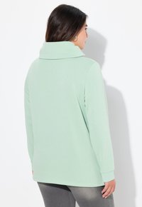 Maglia verde chiaro a maniche lunghe con colletto alto, texture morbida, spalle scese e orlo arrotondato, abbinata a pantaloni grigi scuri slim.