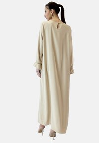 Langes beiges Kleid aus glattem Stoff, mit langen Ärmeln, einem Schlüsselloch-Ausschnitt hinten mit einem Knopf und gerafften Manschetten. Silberne High Heels vervollständigen den Look.