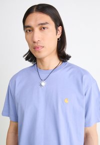 Light purple cotton t-shirt with a crew neck and short sleeves. Features a small yellow logo on the chest and a silver sun pendant necklace.