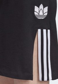 Czarna spódnica z bocznym rozcięciem odsłaniającym nogę, z białym logo trefoil Adidas oraz pionowymi czarno-białymi paskami z tkaniny.