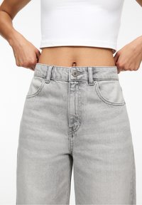 Donna che indossa jeans a vita alta grigi chiari e un top corto bianco, mentre regola la vita con entrambe le mani.