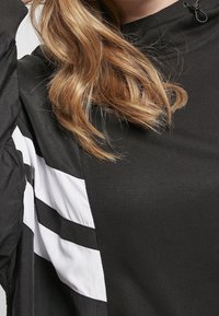 Blond vlasy padají přes černý top s límcem na šňůrku a na levé straně je vidět rukáv větrovky s černobílými pruhy.
