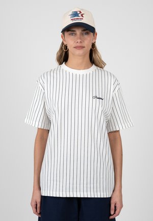 Kvinde iført hvid skjorte med tynde striber og korte ærmer, navy shorts, beige baseballkasket med rød og blå logo samt guldringe i ørerne, stående mod en ensfarvet baggrund.