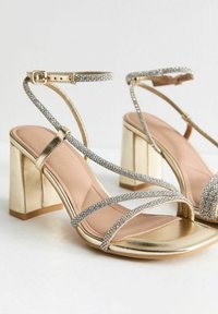 Gold high-heeled sandals featuring a block heel, multiple thin straps with rhinestones, and a smooth metallic finish on the sole.
