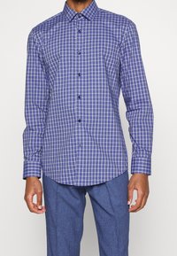 Camicia a maniche lunghe a quadretti blu e bianchi con colletto a bottoni e bottoni neri. Presenta un orlo curvo e polsini aderenti.