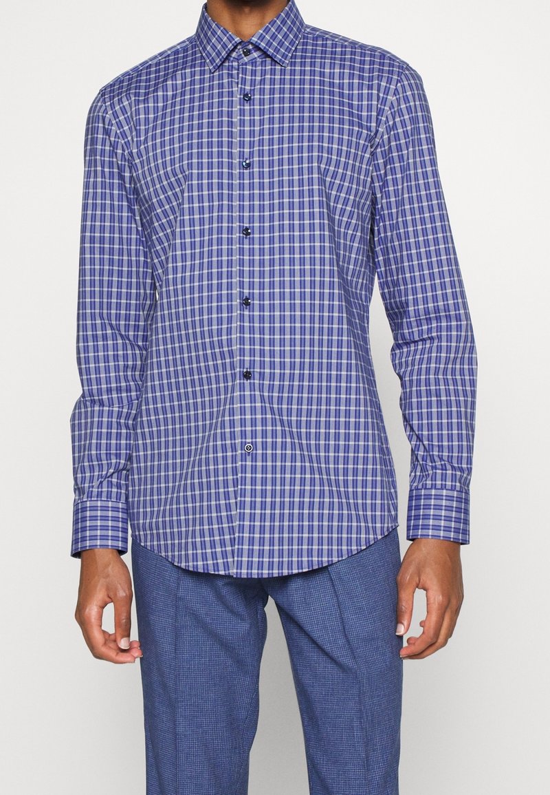 Camicia a maniche lunghe a quadretti blu e bianchi con colletto a bottoni e bottoni neri. Presenta un orlo curvo e polsini aderenti.