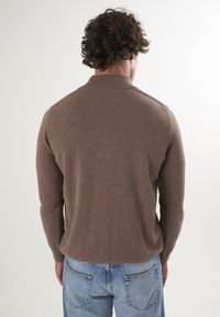 Brązowy sweter z mieszanki bawełny z długimi rękawami i ribbowym kołnierzykiem, noszony z jasnoniebieskimi, postarzanymi dżinsami, widok z tyłu.