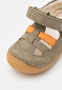 Sandalo per bambini in suede grigio con accenti arancioni, punta chiusa, lati ritagliati e strap in velcro. Suola in gomma con dettagli di cucitura.