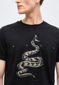 Άνδρας με μαύρο μπλουζάκι που φέρει μεγάλο γραφικό ενός κουλουριασμένου οχιάς με το κεφάλι ανασηκωμένο και το στόμα ανοιχτό στην περιοχή του στήθους.