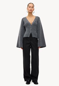 Grijze wollen cardigan met een diepe V-hals, knopen aan de voorkant en uitlopende mouwen, gecombineerd met zwarte wijde broek en zwarte schoenen.