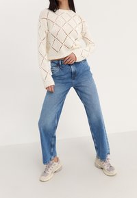 Gebreide crème trui met ruitvormige uitsparingen, gecombineerd met lichtblauwe high-waisted jeans en beige sneakers. Ontspannen pasvorm en wijde pijpen.