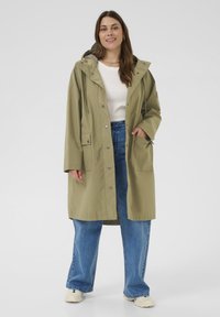 Veste de pluie vert olive avec capuche, fermeture à pression à l'avant, deux poches latérales et cordons ajustables, portée par-dessus un haut blanc et un jean bleu.