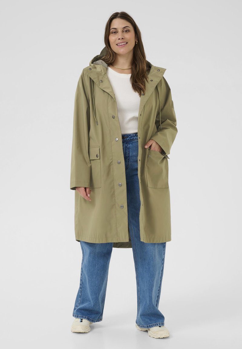 Veste de pluie vert olive avec capuche, fermeture à pression à l'avant, deux poches latérales et cordons ajustables, portée par-dessus un haut blanc et un jean bleu.