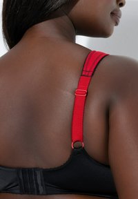 Sort bh med en tekstureret finish, der har røde justerbare stropper og lukning med krog. Stroppen har en dekorativ kantdetalje.