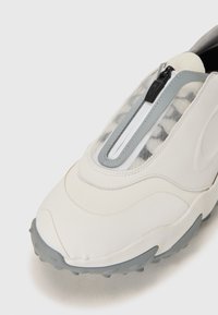Baltas slip-on sporta apavi ar teksturētu pelēku zoli, ar caurspīdīgu acu paneli un vidusdaļas rāvējslēdzēju. Gluda sintētiska materiāla.