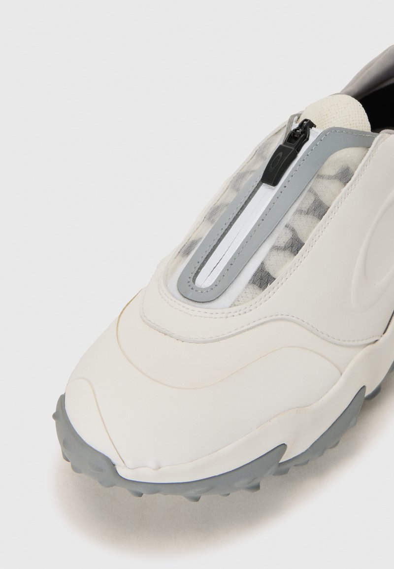 White slip-on sneaker with a textured grey sole, featuring a transparent mesh panel and a central zip closure. Smooth synthetic material.