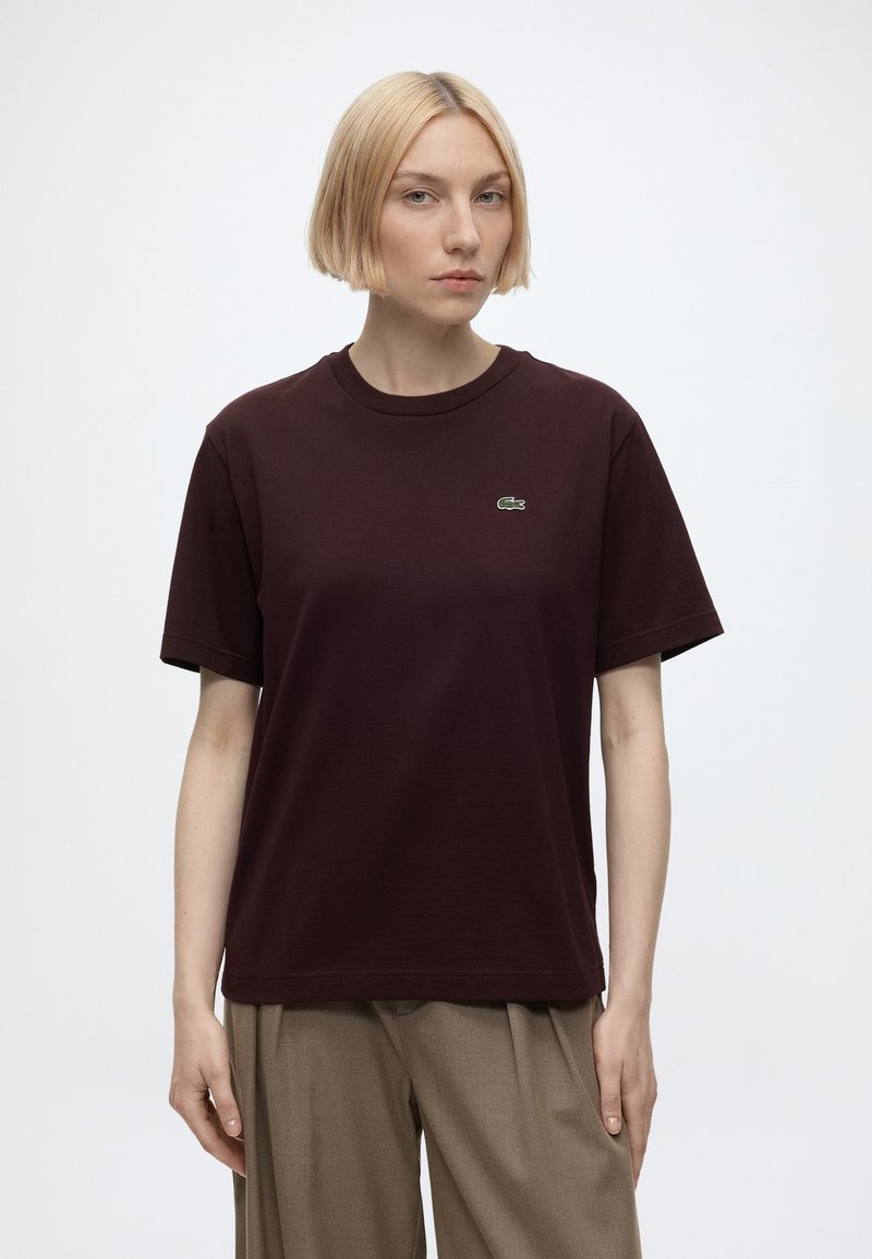 Femme blonde portant un t-shirt marron foncé à manches courtes avec un petit logo crocodile vert sur la poitrine et un pantalon beige ample.