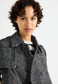Grå herringbone trenchcoat med utskjært krage, to store knapper og en løs passform, brukt over en svart og hvit stripet topp.
