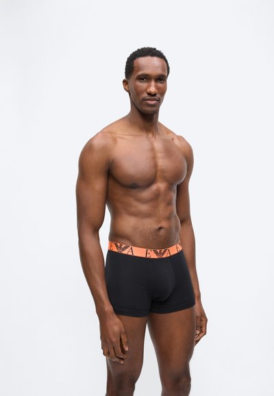 Μυώδης άνδρας που στέκεται με μαύρα μποξεράκια με πορτοκαλί ζώνη μέσης που φέρει λογότυπο, πάνω σε μια απλή ανοιχτή επιφάνεια φόντου.