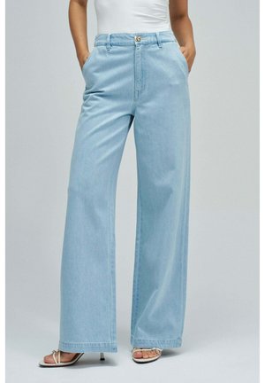 Modell trägt hellblaue Jeans mit weitem Bein, Vordertaschen und Knopfverschluss, kombiniert mit weißen, geschnürten Sandalen mit Absatz.