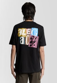 Melnā īsroka T-krekls ar daudzkrāsainu "LEGACY" grafiku uz muguras, katram burtam ar dažādiem faktūras rakstiem un stiliem.