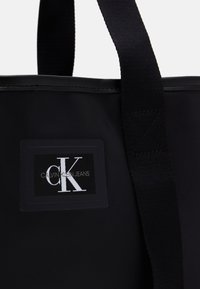 Black tote bag featuring fabric material, prominent logo patch with "Calvin Klein Jeans," and black woven handles. Smooth texture.