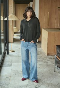 Femme se tenant à l'intérieur, portant un haut à manches longues gris foncé, un jean léger à jambes larges bleu clair et des chaussures rouges, les mains dans les poches.