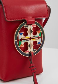 Rød læderhåndtaske med en cirkulær guldlås med flerfarvede geometriske indlæg og en bindesnor. Glat tekstur med struktureret design.