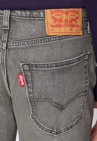 Vaqueros de mezclilla gris con un diseño de bolsillo trasero distintivo, una etiqueta roja de Levi's y un parche de cuero amarillo con logotipos y detalles.