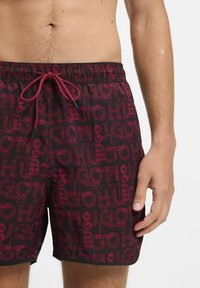 Punaiset ja mustat uimashortsit, joissa on joustava vyötärö ja naru. Ominaisuuksiin kuuluu toistuva 'HUGO' -logo eri kokoisin ja läpinäkyvinä.