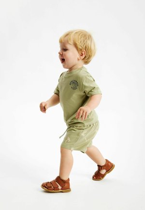 Tricou din bumbac verde cu mâneci scurte și text imprimat, combinat cu șorturi cu șnur asortate și sandale maronii cu mai multe curele.