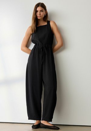 Young woman standing against white wall wearing sleeveless black jumpsuit with drawstring waist and black mesh flats.