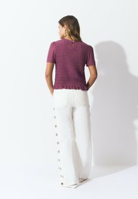 Haut smocké violet avec des manches courtes bouffantes, doté d'une fermeture arrière à boutons. Associé à un pantalon large blanc orné de boutons sur les côtés.
