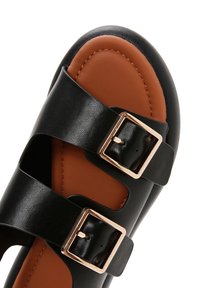 Black leather sandal with two wide straps and gold buckles, featuring a brown cushioned footbed. Smooth texture and minimal design.