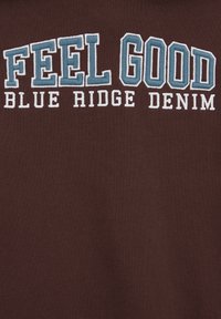 Teksti "HYVÄ TUNTEA" suurilla sinisillä kirjaimilla ja "BLUE RIDGE DENIM" pienemmillä valkoisilla kirjaimilla ruskealla kankaasta taustalla.