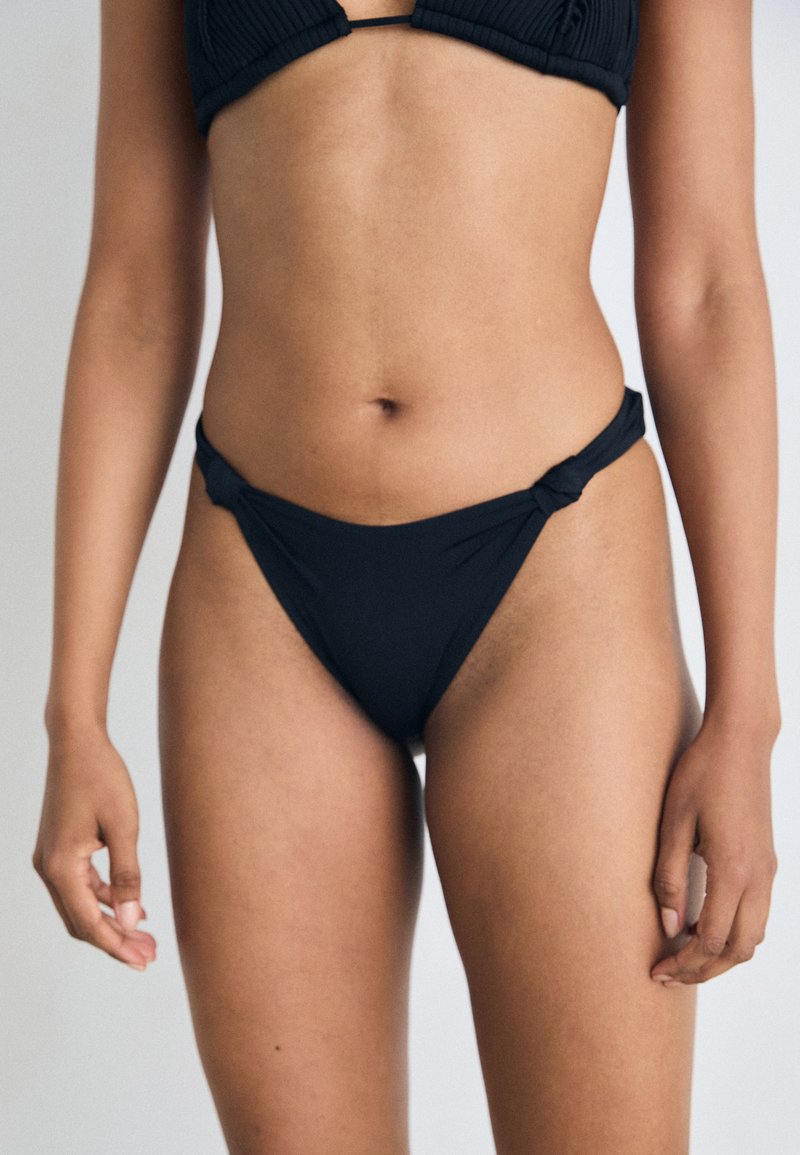 Black bikini top with ribbed texture and a tied knotted detail. The matching bottom features a high-cut design with minimal coverage and smooth finish.