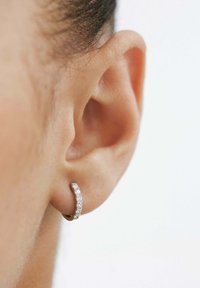 Pendiente de aro de plata con pequeños cristales transparentes incrustados, exhibido contra una piel suave y cabello oscuro, destacando su diseño curvado.