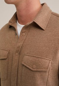 Ruda mygtukų marškiniai, pagaminti iš minkštos, tekstūruotos medžiagos, su dviem krūtinės kišenėmis ir apykakle; turi apvalius rudus snap mygtukus.